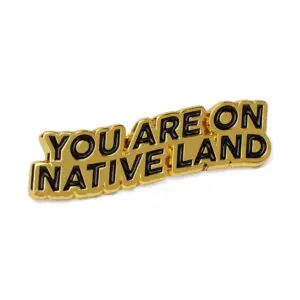 Gold native land pin featuring 'You Are on Native Land' text, shown on a denim jacket